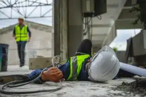 A construction worker lying on the floor with a serious injury after falling over. Another worker runs towards them to help.