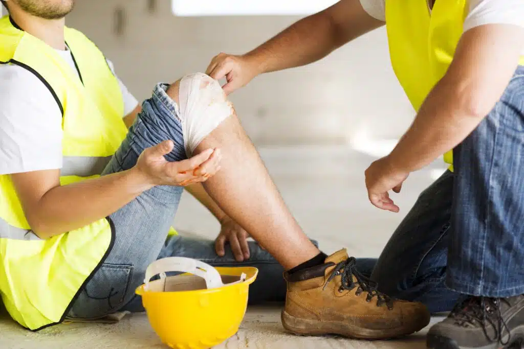 a construction workers in a high vis jacket receiving first aid from a co-worker after a soft tissue injury to the knee